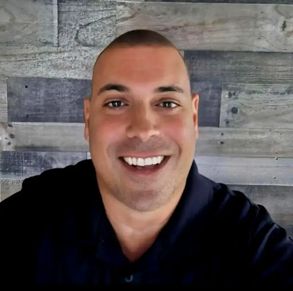 Bald man with a wide smile, wearing a dark polo shirt against a wood-grain backdrop.