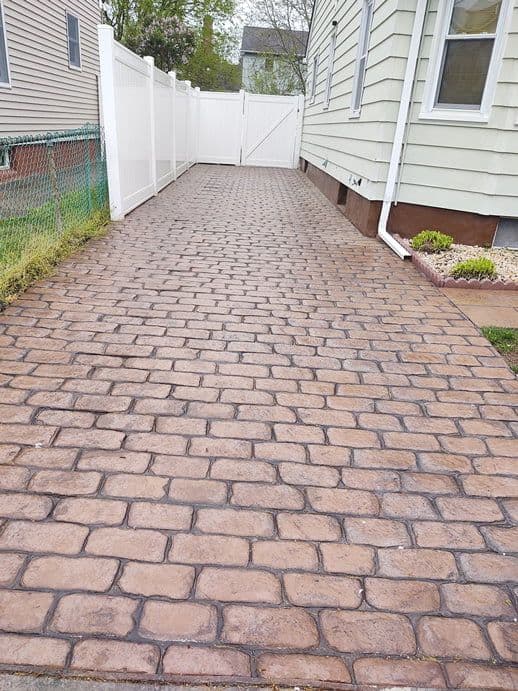 A stamped concrete path in a brick pattern running between a house and a fence